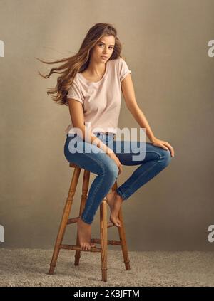 Geht hin, Mädchen. Studioaufnahme einer schönen jungen Frau, die auf einem Hocker vor einem schlichten Hintergrund sitzt. Stockfoto