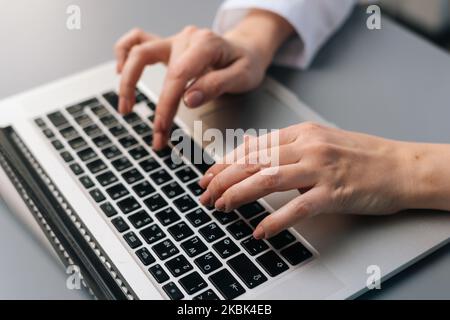Nahaufnahme aus einem hohen Winkel der nicht erkennbaren weiblichen Ärztin in weißer medizinischer Uniform, die auf der Laptop-Tastatur am Schreibtisch sitzt. Stockfoto