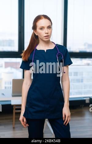 Vertikales Porträt einer ernsthaften Rotschopf-Ärztin, die eine dunkle medizinische Uniform trägt und die Kamera auf dem Hintergrund des Fensters betrachtet. Stockfoto