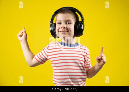 Fröhlicher Junge mit Kopfhörern und T-Shirt zeigt seine Finger auf einen gelben Hintergrund Stockfoto