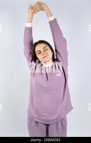 Junge Brünette Frau in Musselin Lavendel Schlafanzug Schlafanzug erstreckt sich, indem sie ihre Arme hochhebt und ihre Augen schließt Stockfoto