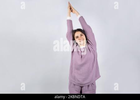 Junge Brünette Frau in Musselin Lavendel Schlafanzug Schlafanzug erstreckt sich, indem sie ihre Arme nach oben. Stockfoto