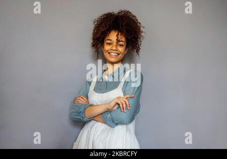 Junge schwarze Frau trägt beige Schürze isoliert auf grauem Hintergrund mit Kopieplatz. Porträt einer erfolgreichen afroamerikanischen Frau an der grauen Wand. Lächelnd Stockfoto