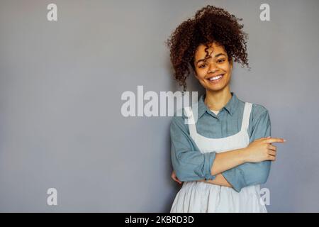 Junge schwarze Frau trägt beige Schürze isoliert auf grauem Hintergrund mit Kopieplatz. Porträt einer erfolgreichen afroamerikanischen Frau an der grauen Wand. Lächelnd Stockfoto