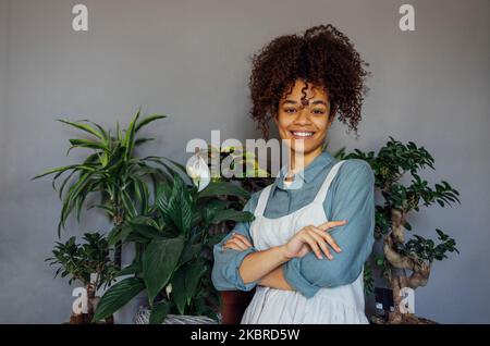 Junge schwarze Frau trägt beige Schürze isoliert auf grauem Hintergrund mit Kopieplatz. Porträt einer erfolgreichen afroamerikanischen Frau mit Pflanzen. Lächelnd Stockfoto