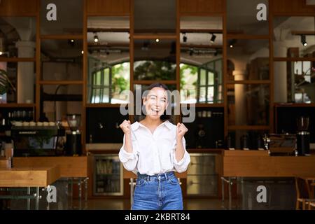 Glückliches, enthusiastisches asiatisches Mädchen, die Geschäftsbesitzerin triumphiert, eröffnet ihr eigenes Restaurant und steht vor einem leeren Café Stockfoto