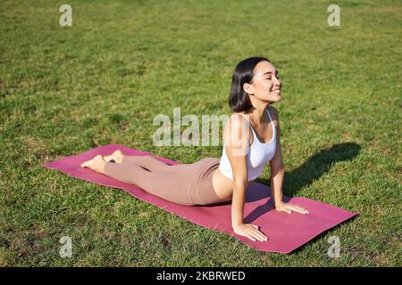 Lächelnde junge Sportlerin streckt sich im Park auf einer Gummimatte, macht Yoga-Asana-Übungen, trainiert an der frischen Luft in Fitnesskleidung Stockfoto