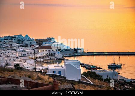 Verträumter Sonnenuntergang über Mykonos Stadt und dem kleinen Hafen während der goldenen Stunde vor der Dämmerung mit dem Himmel, der rot, orange und warme Farben wechselt, wenn die Sonne hinter der Ägäis über Mykonos Stadt oder Chora, auf Myconos Insel in den Kykladen, Ägäis in Griechenland untergeht. Die berühmte griechische Mittelmeerinsel wird mit weiß getünchten traditionellen Gebäuden wie Windmühlen oder einer kleinen Kirche als Insel der Winde bezeichnet. Mykonos ist eine beliebte Insel für Prominente und Touristen, die im pulsierenden Nachtleben feiern möchten. Die Insel gilt auch als homosexuell freundlich. Die griechische Regierung hat den Sommer wieder belebt Stockfoto