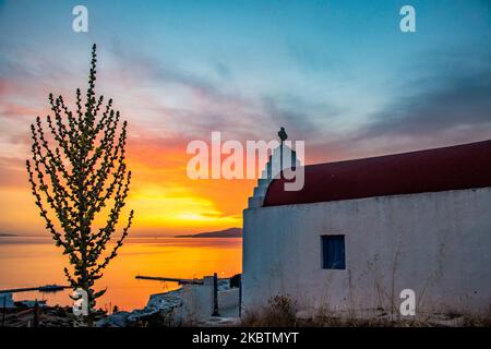 Verträumter Sonnenuntergang während der goldenen Stunde mit einer kleinen traditionellen Kapelle kurz vor der Dämmerung mit dem Himmel in Rot, Orange und warmen Farben, wenn die Sonne hinter der Ägäis über Mykonos Stadt oder Chora untergeht, auf Myconos Insel in den Kykladen, Ägäis in Griechenland. Die berühmte griechische Mittelmeerinsel wird mit weiß getünchten traditionellen Gebäuden wie Windmühlen oder einer kleinen Kirche als Insel der Winde bezeichnet. Mykonos ist eine beliebte Insel für Prominente und Touristen, die im pulsierenden Nachtleben feiern möchten. Die Insel gilt auch als homosexuell freundlich. Die griechische Regierung hat den Sommertouristen wieder belebt Stockfoto