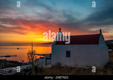 Verträumter Sonnenuntergang während der goldenen Stunde mit einer kleinen traditionellen Kapelle kurz vor der Dämmerung mit dem Himmel in Rot, Orange und warmen Farben, wenn die Sonne hinter der Ägäis über Mykonos Stadt oder Chora untergeht, auf Myconos Insel in den Kykladen, Ägäis in Griechenland. Die berühmte griechische Mittelmeerinsel wird mit weiß getünchten traditionellen Gebäuden wie Windmühlen oder einer kleinen Kirche als Insel der Winde bezeichnet. Mykonos ist eine beliebte Insel für Prominente und Touristen, die im pulsierenden Nachtleben feiern möchten. Die Insel gilt auch als homosexuell freundlich. Die griechische Regierung hat den Sommertouristen wieder belebt Stockfoto