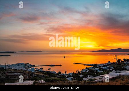 Verträumter Sonnenuntergang über Mykonos Stadt und dem kleinen Hafen während der goldenen Stunde vor der Dämmerung mit dem Himmel, der rot, orange und warme Farben wechselt, wenn die Sonne hinter der Ägäis über Mykonos Stadt oder Chora, auf Myconos Insel in den Kykladen, Ägäis in Griechenland untergeht. Die berühmte griechische Mittelmeerinsel wird mit weiß getünchten traditionellen Gebäuden wie Windmühlen oder einer kleinen Kirche als Insel der Winde bezeichnet. Mykonos ist eine beliebte Insel für Prominente und Touristen, die im pulsierenden Nachtleben feiern möchten. Die Insel gilt auch als homosexuell freundlich. Die griechische Regierung hat den Sommer wieder belebt Stockfoto