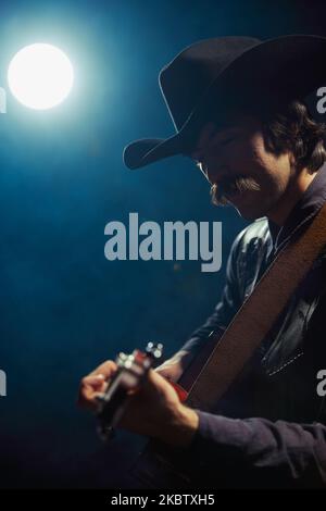 Porträt eines Mannes mit Schnurrbart in Kleidung im Landhausstil, der unter einem Scheinwerfer Gitarre spielt, isoliert auf dunkelblauem Hintergrund mit Rauch Stockfoto