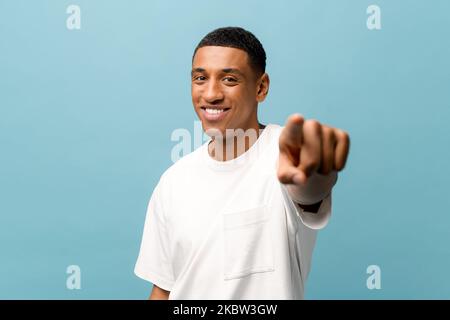 Attraktiver, fröhlicher junger afroamerikanischer Kerl, der ein weißes, legeres T-Shirt trägt, zeigt mit dem Zeigefinger auf die Kamera und wählt Sie isoliert auf blauem Hintergrund aus, glücklicher, tausendjähriger Mann, der die Richtung vor der Kamera zeigt Stockfoto