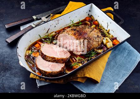 Traditioneller deutscher Schweinebraten, serviert mit Gemüse und Kräutern in dunkler Biersauce, serviert aus nächster Nähe in einer rustikalen Schmorpfanne Stockfoto