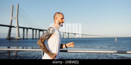 Tätowierte junge Mann beim Joggen an der Flussküste bei Sonnenuntergang. Banner mit einem sportlichen Kerl, der im Freien Cardio-Training macht Stockfoto