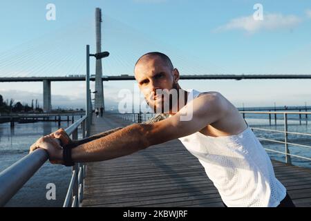 Zielstrebig tätowiert bärtiger junger Mann, der draußen arbeitet. Sportlicher Typ, der die Kamera anschaut, bevor er in der Stadt joggt, moderne Stadtbrücke im Hintergrund Stockfoto