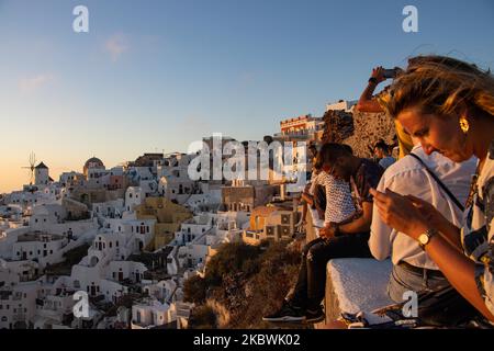 Touristen genießen den atemberaubenden Sonnenuntergang in Oia auf der Insel Santorini, auf den Kykladen, der Ägäis, in Griechenland. Sie befinden sich an einem Aussichtspunkt auf der alten traditionellen Burg und den Dächern von Häusern, Kirchen und Hotels, die auf den vulkanischen Klippen errichtet wurden. Touristen aus der ganzen Welt sind entspannt und fotografieren und Selfies mit Handys und Kameras die Sonne geht im Mittelmeer unter. Die Insel ist berühmt für den magischen Sonnenuntergang, die Architektur der weiß getünchten Häuser, Hotels mit Blick auf den Pool und die blauen Kuppelkirchen am steilen Rand der Klippe. Santorini ist ein beliebtes Ziel für Flitterwochen Stockfoto