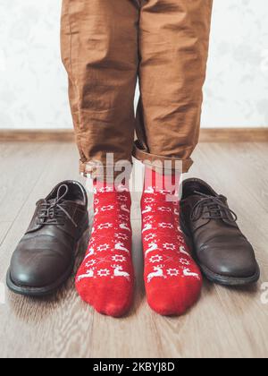 Der junge Mann in Chinohosen und leuchtend roten Socken mit Rentieren ist bereit, Wildlederschuhe zu tragen. Skandinavisches Muster. Winterurlaub. Stockfoto