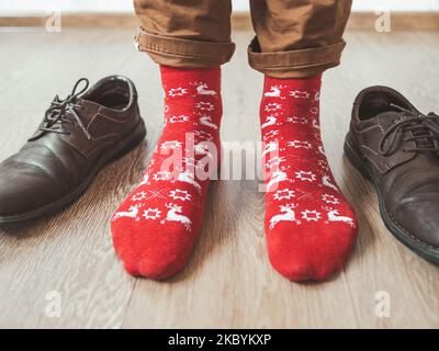 Der junge Mann in Chinohosen und leuchtend roten Socken mit Rentieren ist bereit, Wildlederschuhe zu tragen. Skandinavisches Muster. Winterurlaub. Stockfoto