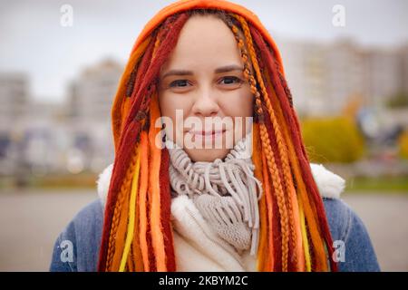 Porträt einer positiven Frau mit hellen Dreadlocks auf der Stadtstraße. Fröhliche Hündin mit bunten Frisuren, die in die Kamera schauen und im trüben Wetter lächeln Stockfoto