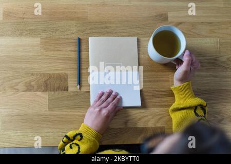 Frau öffnet ein geschlossenes Notizbuch und hält einen weißen Becher mit Tee Stockfoto