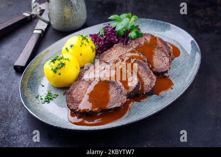 Traditionell geschmorte, marinierte deutsche Sauerbraten aus Rindfleisch mit blauem Kraut und Kartoffelknödeln in würziger dunkler Biersauce, die als Nahaufnahme in einem Nordi serviert werden Stockfoto