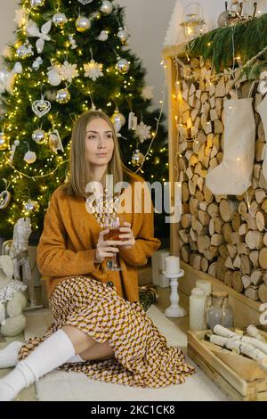 Weihnachtszeit. In Gedanken verloren. Junge schöne verträumte Frau, die in der Nähe des weihnachtsbaums sitzt und an etwas denkt. H Stockfoto
