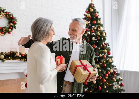 Fröhliche reife Frau tauscht weihnachtsgeschenke mit glücklichem Ehemann in der Nähe verzierte Tanne aus Stockfoto