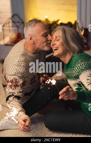 Positiver Mann mittleren Alters, der seine Frau küsst und am heiligabend glänzende Wunderkerzen hält Stockfoto