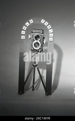 1950s, historisch, eine Doppelreflexkamera, ein Rolleicord, saß auf einem Stativ. Die Worte 'World Famble' - Scramble Letters - sind auf einer Metallumrandung fixiert. Die Rolleicord, eine leichte Doppelreflexkamera im Mittelformat, war eine einfachere, preiswertere Version der professionellen Rolleiflex-Filmkamera und wurde von 1933 bis 1976 in verschiedenen Modellen gefertigt. Stockfoto