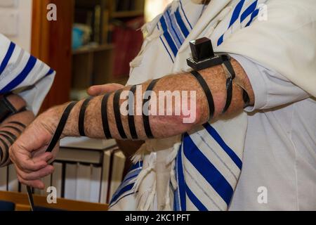 Ein erwachsener Mann, der ein jüdisches Tefillin auf seinen Arm legte und zum Beten einen Gebetsschal trug. Vorbereitung auf ein Gebet Stockfoto