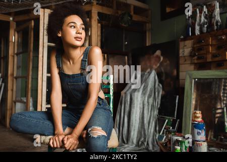 Junge afroamerikanische Frau in Denim-Overalls, die in einem Kunstworkshop in der Nähe von Dosen mit Farbe wegschaut Stockfoto