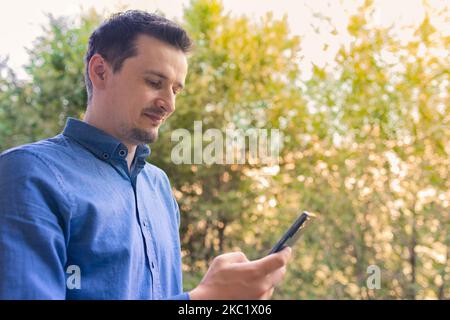 Glücklicher Mann, der im Herbstpark spazierengeht und lächelt. Junger Mann, der auf den Bildschirm des Mobiltelefons schaut und gerne Nachrichten liest. Stockfoto