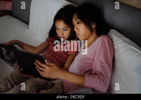 Asiatische Frau und ihre Tochter verwenden Tablet-Computer, während sie zu Hause auf dem Bett sitzen Stockfoto