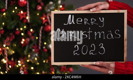 Marry Weihnachtsbanner Konzept. Weibliche Hände in rotem Pullover halten Kreidetafel mit Text Frohe Weihnachten 2023 auf dem Hintergrund von schönen festlichen Christm Stockfoto