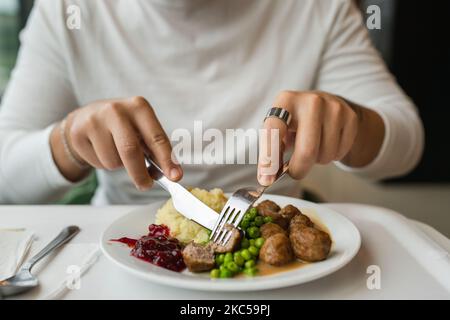 Nahaufnahme der Hände eines unbekannten kaukasischen Mannes, der mit einer Gabel am Tisch sitzt und Fleischbällchen mit Erbsen und Kartoffelpüree isst Stockfoto