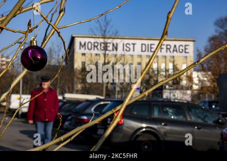 An den Ästen eines jungen, blattlosen Baumes hängt eine zerbrochene Weihnachtskugel, während am 8. Dezember 2020 an der Fassade des Techno-Clubs Berghain ein Transparent mit der Aufschrift „Morgen ist die Frage“ von Künstler Rirkrit Tiravanija hängt. Deutschland, wenn es vor einer Weihnachtssaison mit restriktiven Regeln steht, die private Versammlungen, Handel, Bewegung usw. einschränken, sich von den Bundes- und Kommunalbehörden dazu entschlossen hat, auf die zweite Welle der Coronavirus-Pandemie zu reagieren, da die Anzahl der Fälle nach mehr als einem Monat teilweiser Sperrung, Nimmt ab, aber weit von der gewünschten Schwelle von 50 Fällen fo Stockfoto