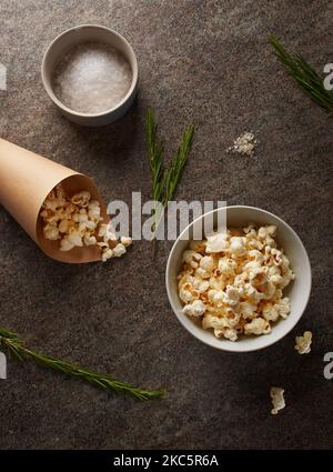 Weiße Porzellanschüssel und brauner Papierkegel voller Popcorn mit Salz und grünen Rosmarinzweigen auf der Marmorplatte Stockfoto