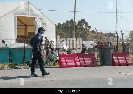 Ein Polizist, der vor dem Neuen provisorischen Flüchtlingslager in Kara Tepe - Mavrovouni patrouilliert, große weiße Zelte mit dem UNHCR- oder UNICEF-Logo hat und Asylbewerber Schlange stehen, um sich anzumelden und Vorräte wie Nahrung, Betten, Kleidung usw. vom Haupteingang aus zu sehen. Das neue eingezäunte Lager ist ein ehemaliges Militärgebiet, Schießplatz der Hellenischen Armee, in der Nähe der Stadt Mytilene auf der Insel Lesbos in Griechenland. Das neue Lager wurde nach dem Brand vom 9. September 2020 gegründet, als eines der größten Flüchtlingslager Europas, das Moria-Flüchtlingslager (Aufnahme-, Identifikations- und Registrierungszentrum), war Stockfoto