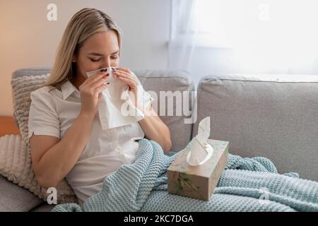 Kranke Frau, die Nase bläst und unter der Decke sitzt. Kranke Frau mit saisonalen Infektionen, Grippe, Allergie im Bett liegend. Kranke Frau mit einer Decke bedeckt Stockfoto