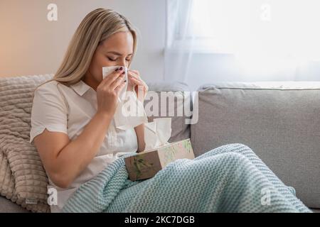 Kranke Frau, die Nase bläst und unter der Decke sitzt. Kranke Frau mit saisonalen Infektionen, Grippe, Allergie im Bett liegend. Kranke Frau mit einer Decke bedeckt Stockfoto
