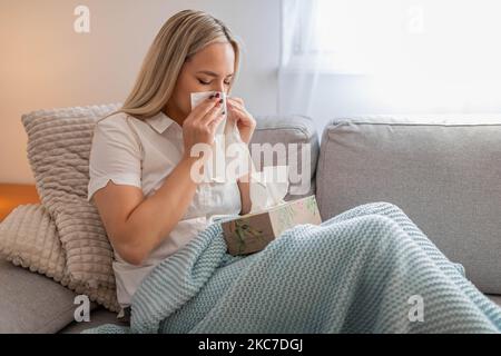 Kranke Frau, die Nase bläst und unter der Decke sitzt. Kranke Frau mit saisonalen Infektionen, Grippe, Allergie im Bett liegend. Kranke Frau mit einer Decke bedeckt Stockfoto