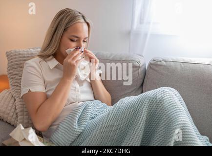 Kranke Frau, die Nase bläst und unter der Decke sitzt. Kranke Frau mit saisonalen Infektionen, Grippe, Allergie im Bett liegend. Kranke Frau mit einer Decke bedeckt Stockfoto