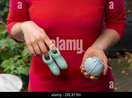 Frau in rotem Kleid mit handgefertigten Häkelbaby Socken oder Pantoffeln aus Wolle in der einen Hand und Ball aus Wolle mit Häkelnadel in der anderen Hand, wählen Stockfoto