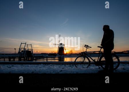 Ein Mann genießt den Sonnenuntergang neben seinem Fahrrad. Zauberhaftes Gestrüppel rund um die Sonnenuntergangszeit und Dämmerung mit klarem Winterhimmel an den gefrorenen Kanälen und Windmühlen von Kinderdijk in den Niederlanden. Das Land ist mit arktischem Wetter, schweren niedrigen Temperaturen, Schneefall und starken Winden aufgrund des Sturms Darcy konfrontiert, der Probleme beim Transport und im täglichen Leben verursachte. Viele der Kanäle wurden eisgefroren, da die Temperatur unter Null liegt, da das kalte Wetter manchmal -16C erreicht, während das Land von Schnee bedeckt ist. Kinderdijk und die Windmühlen im Alblasserwaard Polder, ist eine berühmte Attraktion, ein niederländisches la Stockfoto