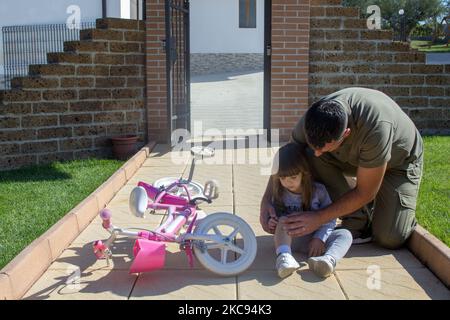 Bild eines jungen Vaters, der seiner Tochter hilft, die auf ihr Fahrrad gefallen ist und ein gehäutes Knie hat. Kleines Mädchen auf der Suche nach Hilfe nach dem Sturz mit ihrem Fahrrad Stockfoto