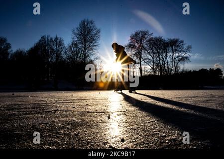 Dunkle Silhouetten von Menschen beim Eislaufen auf dem kristallklaren natürlichen gefrorenen See in der Region Nordbrabant am Nachmittag vor dem Sonnenuntergang. Eislaufrausch übernimmt die Niederlande. Die Niederländer haben viel Spaß und genießen das einwöchige, kalte Schneewetter mit Temperaturen unter Null, die dazu führen, dass Seen, Bäche, Teiche, Flüsse und Kanäle gefrieren. Im Park Meerland in der Nähe der Stadt Eindhoven haben sich die Massen an einem sonnigen Tag mit blauem Himmel abgesetzt, da es eine niederländische Tradition ist. Außerdem werden die Menschen im Freien in der eisgefrorenen Natur und der kalten Umgebung gesehen, die auf dem Eis der gefrorenen Seen spazieren gehen, p Stockfoto