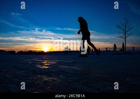 Dunkle Silhouetten von Menschen beim Eislaufen auf dem kristallklaren natürlichen gefrorenen See in der Region Nordbrabant am Nachmittag vor dem Sonnenuntergang. Eislaufrausch übernimmt die Niederlande. Die Niederländer haben viel Spaß und genießen das einwöchige, kalte Schneewetter mit Temperaturen unter Null, die dazu führen, dass Seen, Bäche, Teiche, Flüsse und Kanäle gefrieren. Im Park Meerland in der Nähe der Stadt Eindhoven haben sich die Massen an einem sonnigen Tag mit blauem Himmel abgesetzt, da es eine niederländische Tradition ist. Außerdem werden die Menschen im Freien in der eisgefrorenen Natur und der kalten Umgebung gesehen, die auf dem Eis der gefrorenen Seen spazieren gehen, p Stockfoto