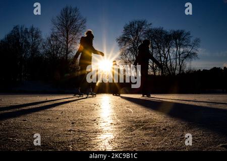 Dunkle Silhouetten von Menschen beim Eislaufen auf dem kristallklaren natürlichen gefrorenen See in der Region Nordbrabant am Nachmittag vor dem Sonnenuntergang. Eislaufrausch übernimmt die Niederlande. Die Niederländer haben viel Spaß und genießen das einwöchige, kalte Schneewetter mit Temperaturen unter Null, die dazu führen, dass Seen, Bäche, Teiche, Flüsse und Kanäle gefrieren. Im Park Meerland in der Nähe der Stadt Eindhoven haben sich die Massen an einem sonnigen Tag mit blauem Himmel abgesetzt, da es eine niederländische Tradition ist. Außerdem werden die Menschen im Freien in der eisgefrorenen Natur und der kalten Umgebung gesehen, die auf dem Eis der gefrorenen Seen spazieren gehen, p Stockfoto