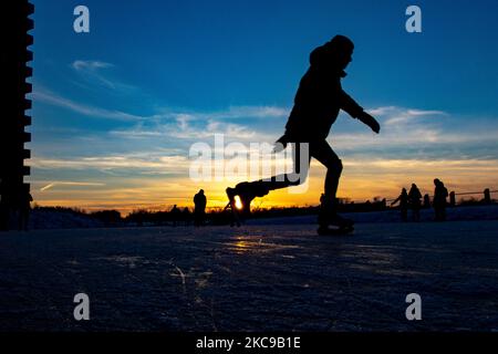 Dunkle Silhouetten von Menschen beim Eislaufen auf dem kristallklaren natürlichen gefrorenen See in der Region Nordbrabant am Nachmittag vor dem Sonnenuntergang. Eislaufrausch übernimmt die Niederlande. Die Niederländer haben viel Spaß und genießen das einwöchige, kalte Schneewetter mit Temperaturen unter Null, die dazu führen, dass Seen, Bäche, Teiche, Flüsse und Kanäle gefrieren. Im Park Meerland in der Nähe der Stadt Eindhoven haben sich die Massen an einem sonnigen Tag mit blauem Himmel abgesetzt, da es eine niederländische Tradition ist. Außerdem werden die Menschen im Freien in der eisgefrorenen Natur und der kalten Umgebung gesehen, die auf dem Eis der gefrorenen Seen spazieren gehen, p Stockfoto